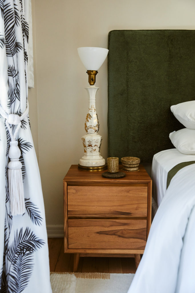 Nightstand, green headboard, white bedding, black and white curtains, white tassel, rug, tan walls, tall lamp, pottery