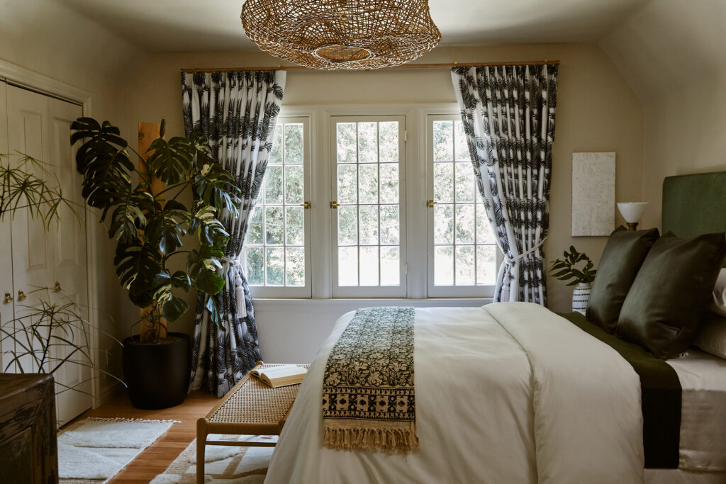 tan walls, queen, bed, green and white blanket, white duvet, black and white curtains, basket, pendant, wood, bench, large tall plant in corner