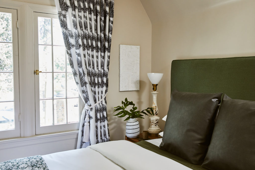 Corner of bedroom, black and white curtains, white bedding, black pillows, green headboard, white painting