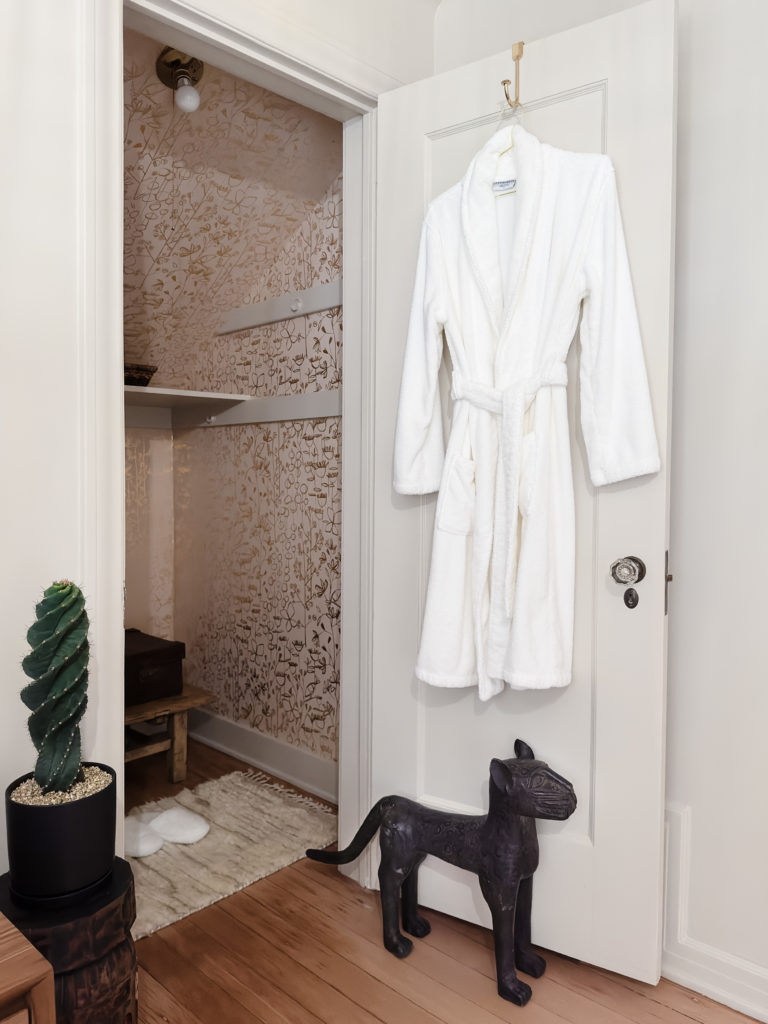 Pink wallpaper, iron, cat doorstop, white robe, hanging off door, spiral cactus in black pot, rug on floor